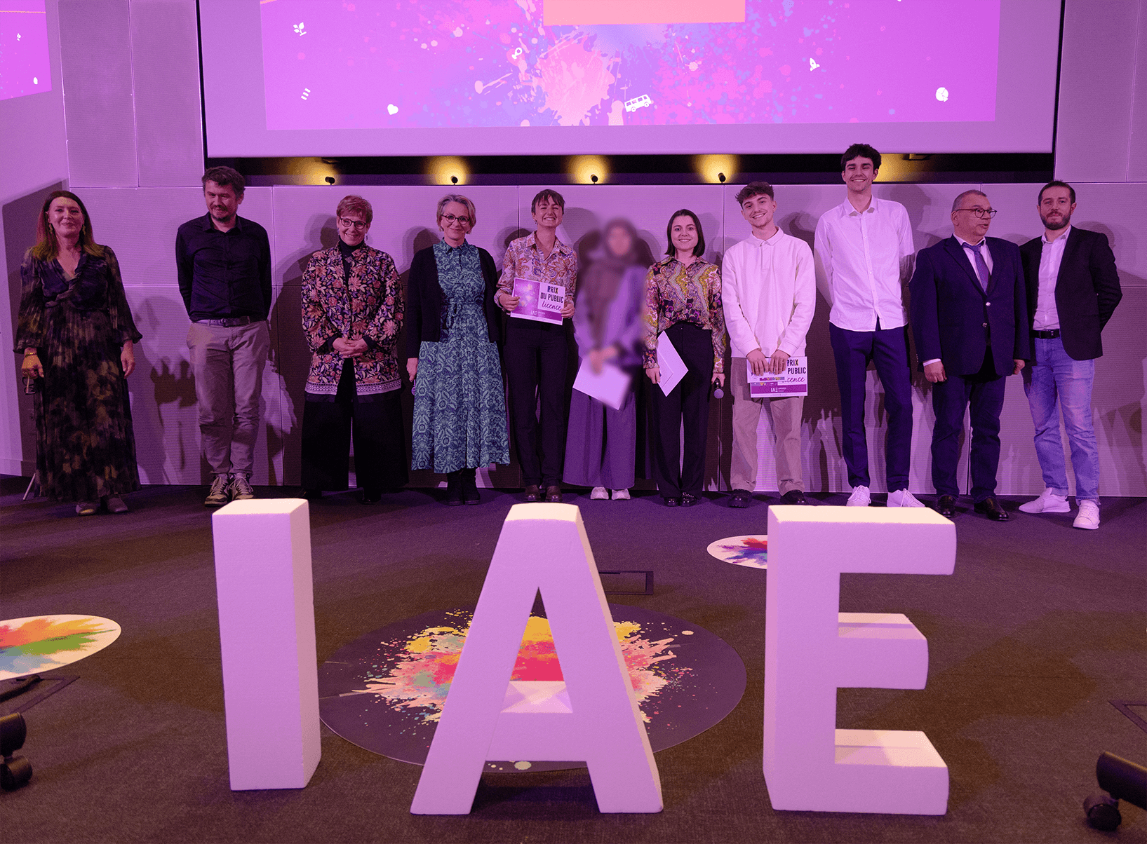 Remise de prix lors de la finale 2025 des Ateliers de l’innovation de l'IAE Limoges. Lauréats et membres du jury sont alignés sur scène, tenant diplômes et trophées, devant de grandes lettres formant « IAE ». L'événement met en lumière les projets innovants des étudiants.
