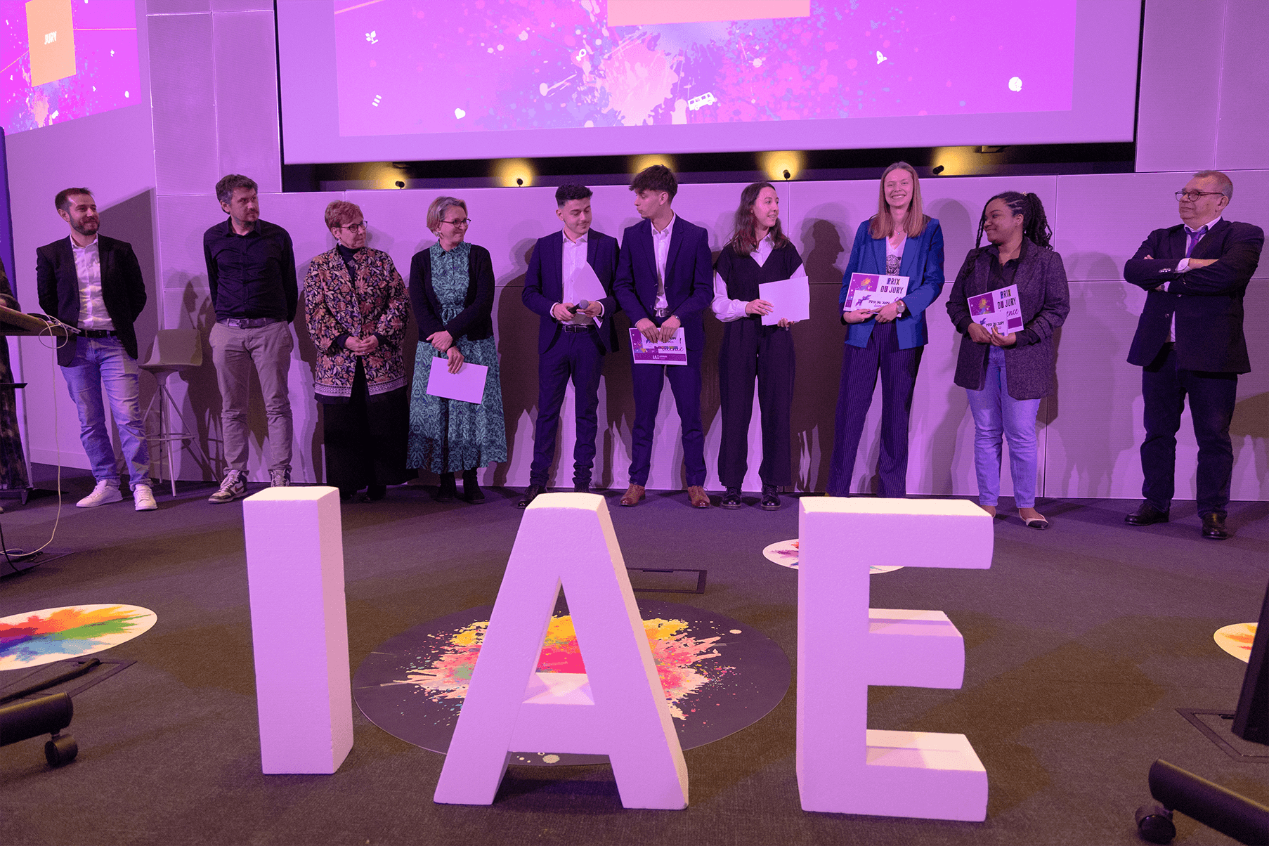 Remise de prix lors de la finale 2025 des Ateliers de l’innovation de l'IAE Limoges. Lauréats et membres du jury sont alignés sur scène, tenant diplômes et trophées, devant de grandes lettres formant « IAE ». L'événement met en lumière les projets innovants des étudiants.