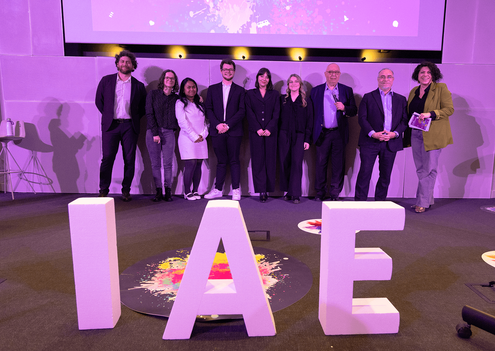Remise de prix lors de la finale 2025 des Ateliers de l’innovation de l'IAE Limoges. Lauréats et membres du jury sont alignés sur scène, tenant diplômes et trophées, devant de grandes lettres formant « IAE ». L'événement met en lumière les projets innovants des étudiants.